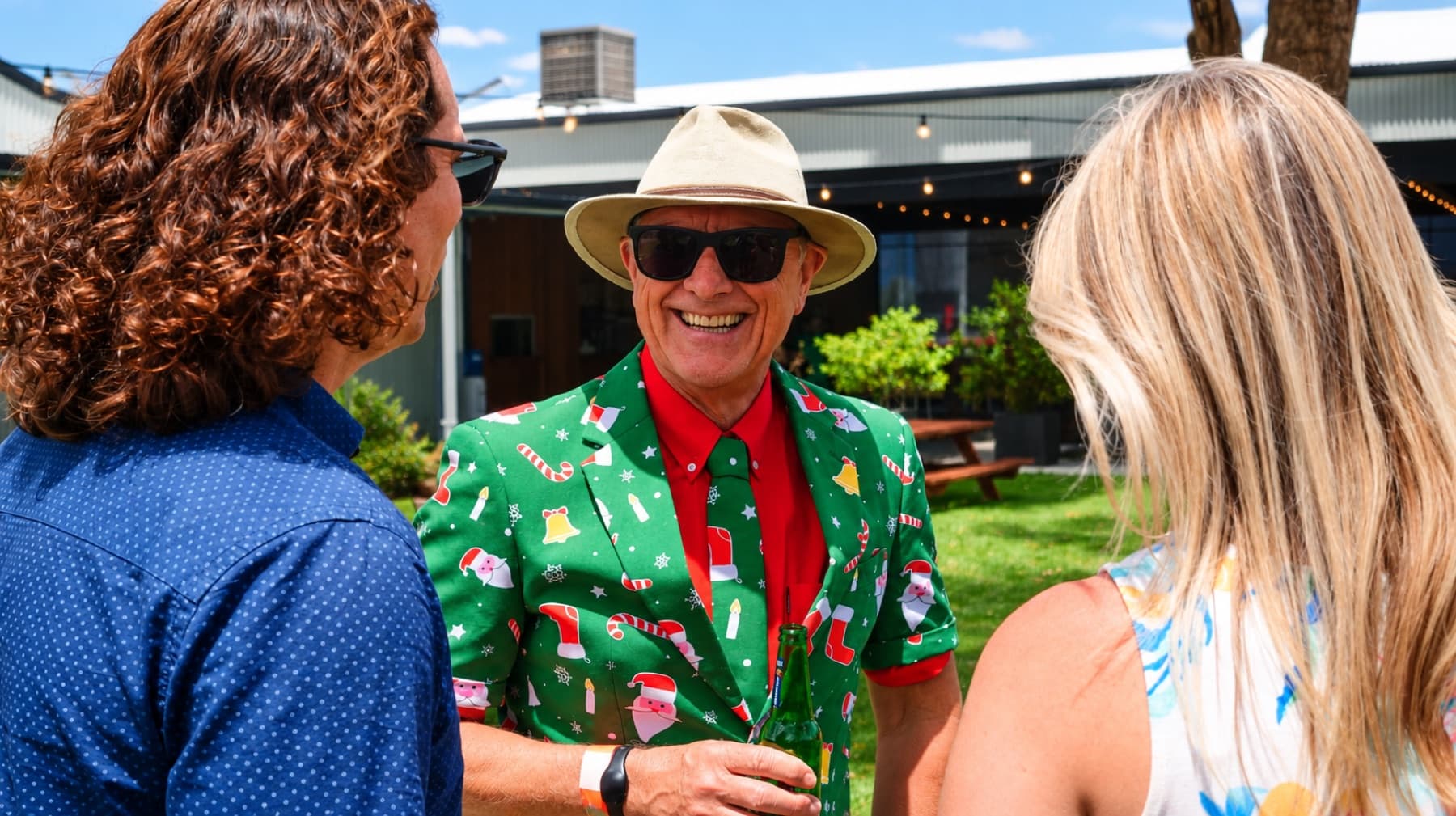 A smiling man in a Christmas-print suit and red tie chats with colleagues at an outdoor end-of-year function. Photo: The Experience Lab