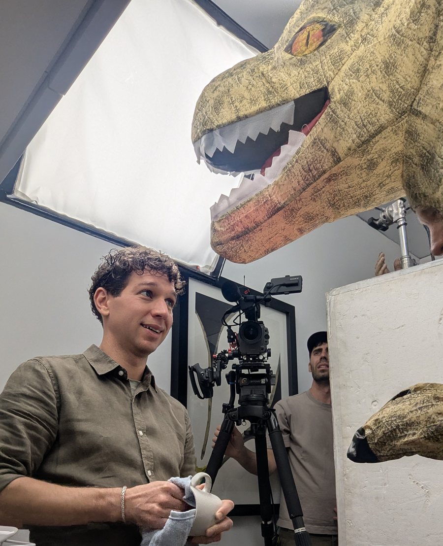 Greg Jarema drying a mug on set, with a crew member behind the camera and a dinosaur costume looming overhead